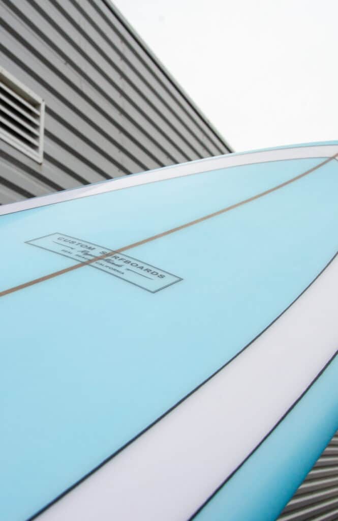 Shapers-Club- Un ROGER HINDS bleu et blanc - TAMAGO - 7'6'' x 22 0 | 22.000'' x 2 7/8'' | 2.875'' - Planche de surf Pailsley adossée à un bâtiment, conçue par Roger Hinds. -surfshop-surfboard