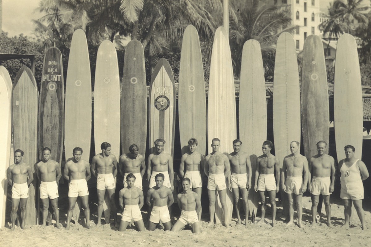 Shapers-Club- Un groupe de surfeurs vintage alignés sur une plage avec leurs longboards, évoquant l'époque classique du surf et reflétant l'histoire des planches de surf. -surfshop-surfboard