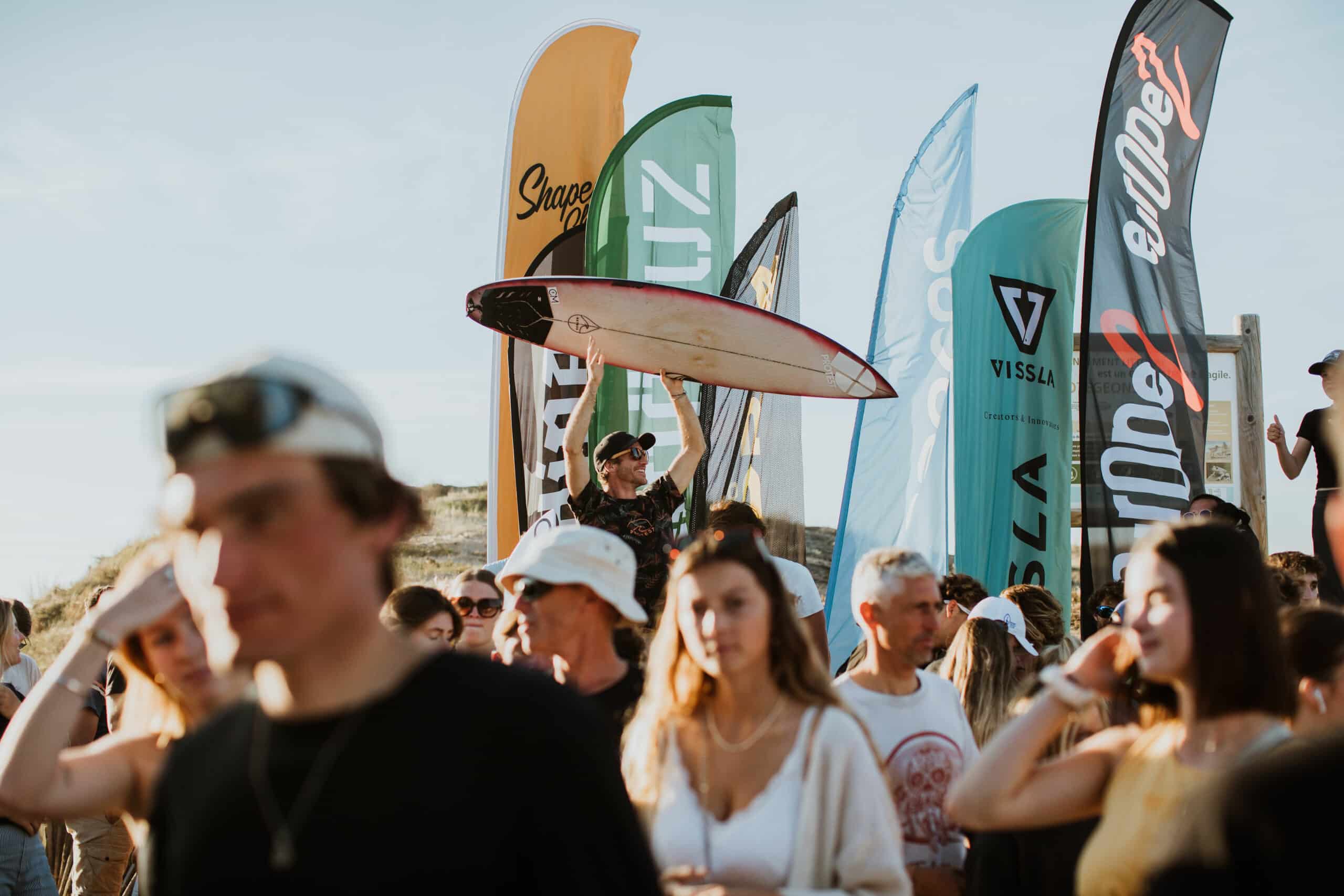 Shapers-Club- Un événement animé en plein air avec une foule de personnes, certaines portant des planches de surf, à proximité de plusieurs banderoles colorées avec des logos de marques sur une plage pour la Qualification Surf Olympique.
 -surfshop-surfboard